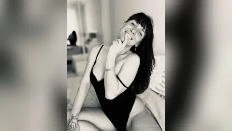 Smiling woman in a black bodysuit relaxing on a bed in a bright, minimalist bedroom setting.
