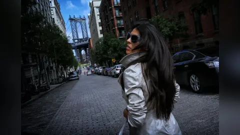 Stylish woman in a white trench coat and sunglasses walking on a cobblestone street near the Manhattan Bridge.