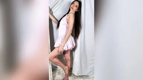 Young woman in a pink satin romper posing against white curtains in a bright, soft-lit room.