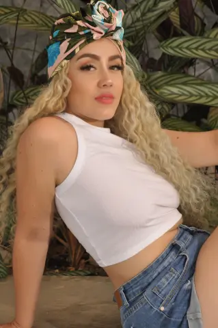 Blonde woman in a white tank top and denim shorts posing outdoors near tropical green plants.
