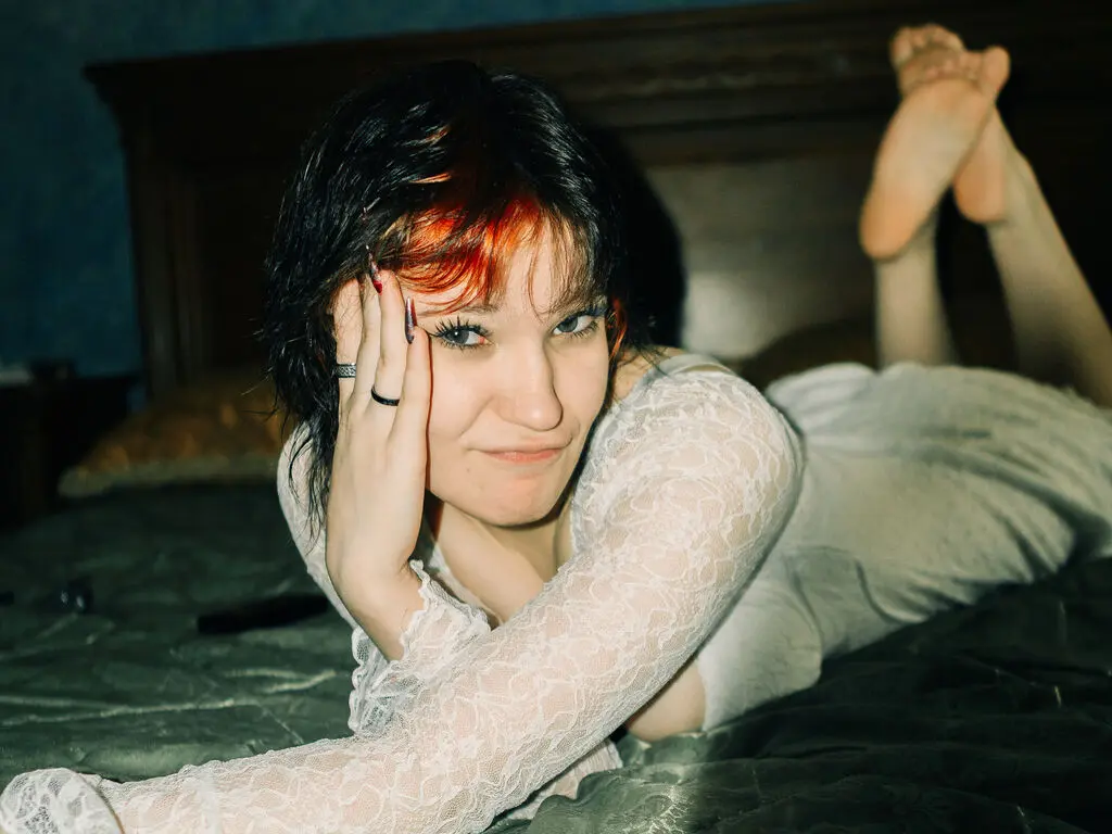 Young woman in a white lace top lying on a bed in a moody, dimly lit bedroom setting.