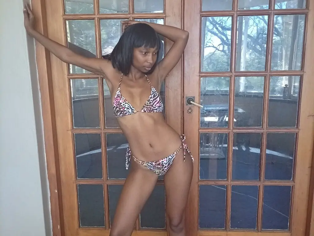 Slim Black woman in a patterned bikini posing confidently by a large wooden glass door in a bright room.