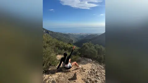 Adventurous woman in black activewear sitting on a rocky mountain peak overlooking a coastal city view.