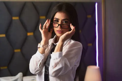 Elegant woman in a white button-down shirt adjusting glasses in a moody, dimly lit bedroom setting.