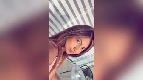 Smiling woman in a pink tank top posing in a bright, modern room with sunlight filtering through blinds.
