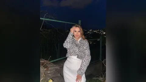 Stylish woman in a leopard print blouse and white skirt posing outdoors at night under city lights.