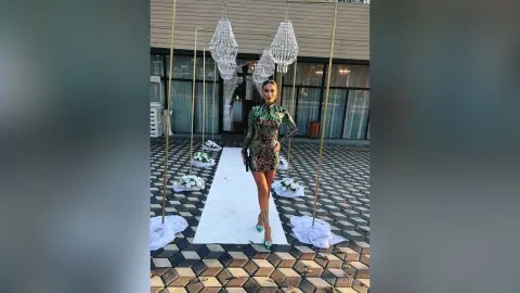 AnnaKarev stands on a white carpet in a tight, sparkly green patterned mini dress and high heels.