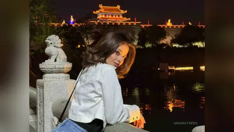 Young woman in a white jacket posing by a stone lion statue at a lit palace at night.
