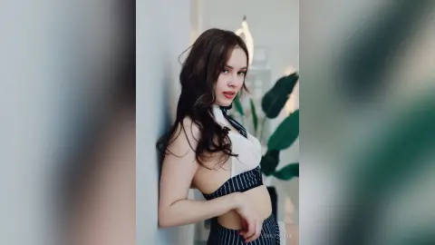 EmilyGiffin poses against a white wall wearing a white and black striped crop top that shows her midriff, with her long dark hair falling over her shoulders.