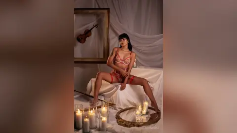 Elegant woman in red lace lingerie sitting on a white bed surrounded by glowing candles in a moody room.