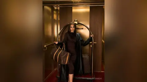 Elegant woman in a black dress holding a designer monogram duffle bag inside a luxury gold elevator.