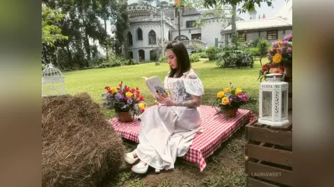 LauriSweet sits on a red and white checkered mat in a garden, wearing a white dress and reading a book outdoors.