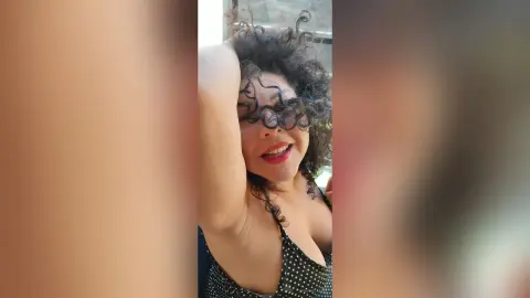 Smiling woman with curly hair in a black polka dot top enjoying bright sunlight indoors.