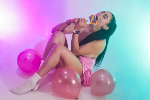 Young woman in pink outfit enjoying a colorful lollipop amidst pink balloons in dreamy neon lighting.