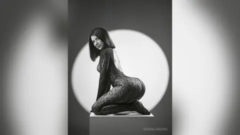 Elegant woman in a black lace bodysuit posing against a circular spotlight in a moody black and white studio.