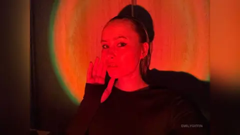 EmilyGiffin poses with one hand near her face, wearing a black long-sleeved shirt against a bright red and orange circular light background.