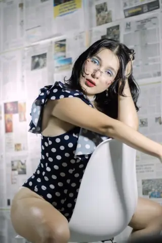 Young woman in a navy polka dot romper posing against a collage newspaper background in a moody studio setting.