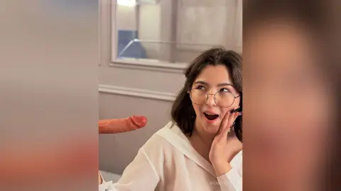 Surprised young woman with glasses in a white blouse laughing in a bright modern indoor setting.