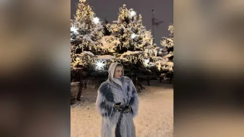 Woman in a cozy grey faux fur coat standing in a snowy forest with twinkling fairy lights at night.