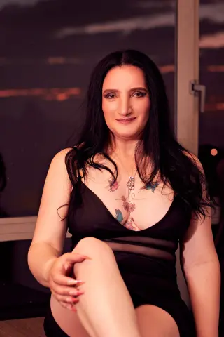 Smiling woman in a black slip dress sitting by a window during a moody evening sunset.