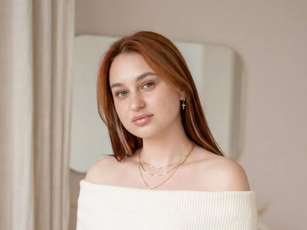 Young woman with reddish hair wearing a white off-the-shoulder top and gold necklace in a bright room.