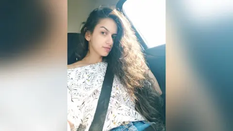 Young woman with long wavy hair wearing a white floral blouse sitting in a car on a bright sunny day.