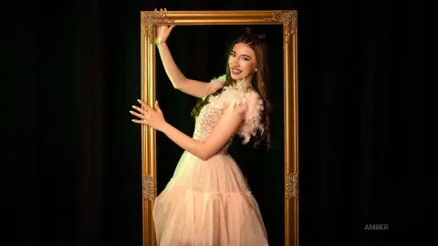 Smiling woman in a glamorous pink feathered gown posing inside a large gold frame against a black backdrop.