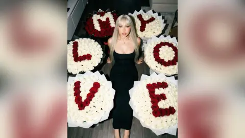 Blonde woman in a sleek black dress sitting amidst large bouquets of red and white roses spelling LOVE.