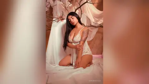 Young woman in white lace lingerie sitting on white sheets in a dreamy, ethereal indoor setting.