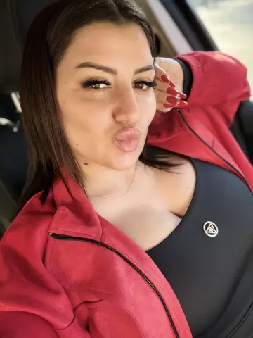 Curvy woman in a red zip-up jacket and black top making a playful pouty face inside a car.