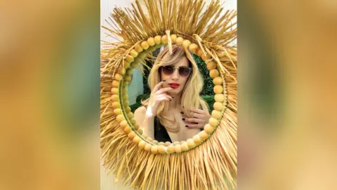 BiankaMoretti poses in a round straw mirror, wearing sunglasses and red lipstick with her blonde hair styled down.