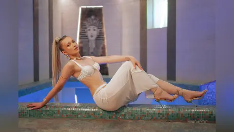 Blonde woman in white bikini and trousers relaxing by a luxury indoor pool with Egyptian decor.