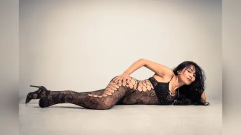 Elegant woman in black lace bodysuit and patterned tights lying on a white floor in a minimalist studio setting.