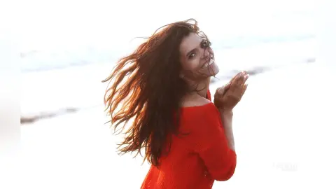 Happy woman with long wavy hair in a red sweater laughing on a bright beach at sunset.