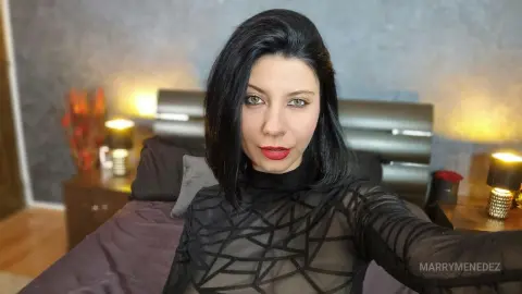 Woman in a black sheer patterned top posing in a cozy bedroom with warm ambient lighting.