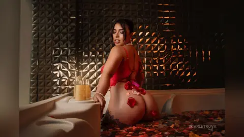 ScarletKova poses naked in a bathtub filled with red rose petals, showing her bare back and buttocks against a textured dark wall.