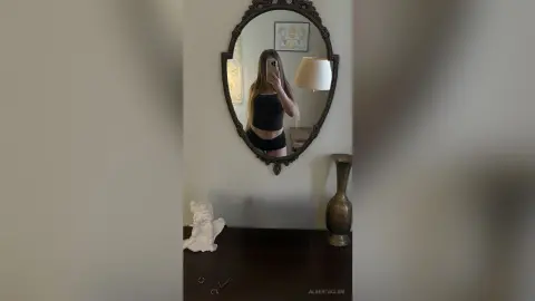 Young woman in a black crop top and shorts taking a mirror selfie in a modern minimalist room.