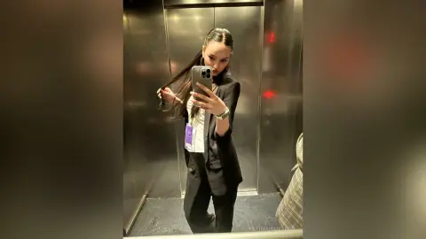 Stylish woman in a black blazer and white shirt taking a mirror selfie in a modern elevator.