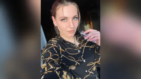 CristaStwell poses in a black button-down shirt with a gold chain pattern, looking at the camera with wet-look hair and light eyes.