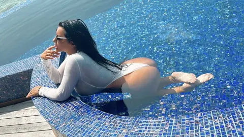 MissyJolie poses in a blue tiled pool wearing a white long-sleeve bodysuit that shows her bare buttocks and legs.