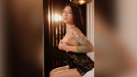 MarlaBolton poses in black lace lingerie, showing off her colorful arm tattoos and long dark hair against a warm, lit background.