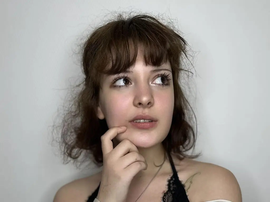 Young woman with brown bangs in a black lace top looking away with a thoughtful expression against a white wall.