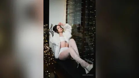 Glamorous woman in a white faux fur coat and sheer tights sitting by a window with twinkling fairy lights.