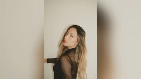 LaurentBelluci poses against a plain wall wearing a sheer black long-sleeve top with her long blonde hair flowing down.