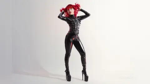 Red-haired woman in a sleek black latex bodysuit and platform heels posing against a white studio background.