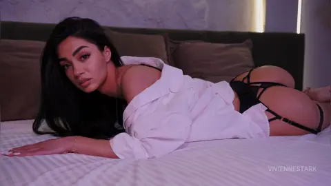 Beautiful woman in white shirt and black lingerie relaxing on a bed in soft, moody lighting.