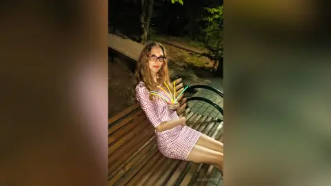 Smiling woman in a red checkered dress sitting on a wooden bench in a dark forest setting.