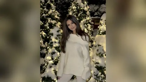 Smiling woman in a cozy white sweater standing amidst glowing snowy Christmas trees at night.