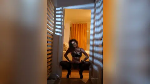 FelishaRose squats low in black lace lingerie, looking down while framed by white slatted blinds.