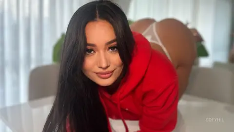 Smiling brunette woman in a red hoodie leaning on a white table in a bright, minimalist room.